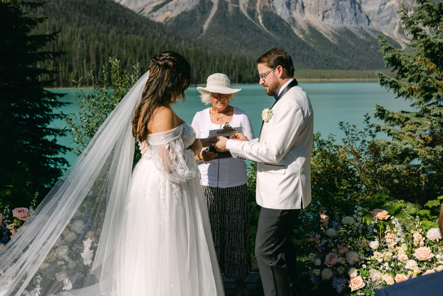 Bride and groom exchanging vows with officiant by a turquoise lake with mountains in the background.