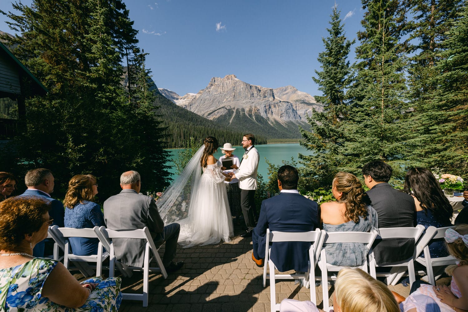 Bride and groom exchanging vows at an outdoor wedding with guests and a scenic mountain lake backdrop.