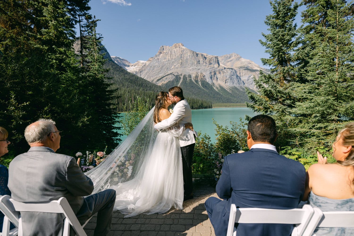 A bride and groom kissing during a wedding ceremony by a turquoise mountain lake, with guests and scenic views of forested mountains in the background.