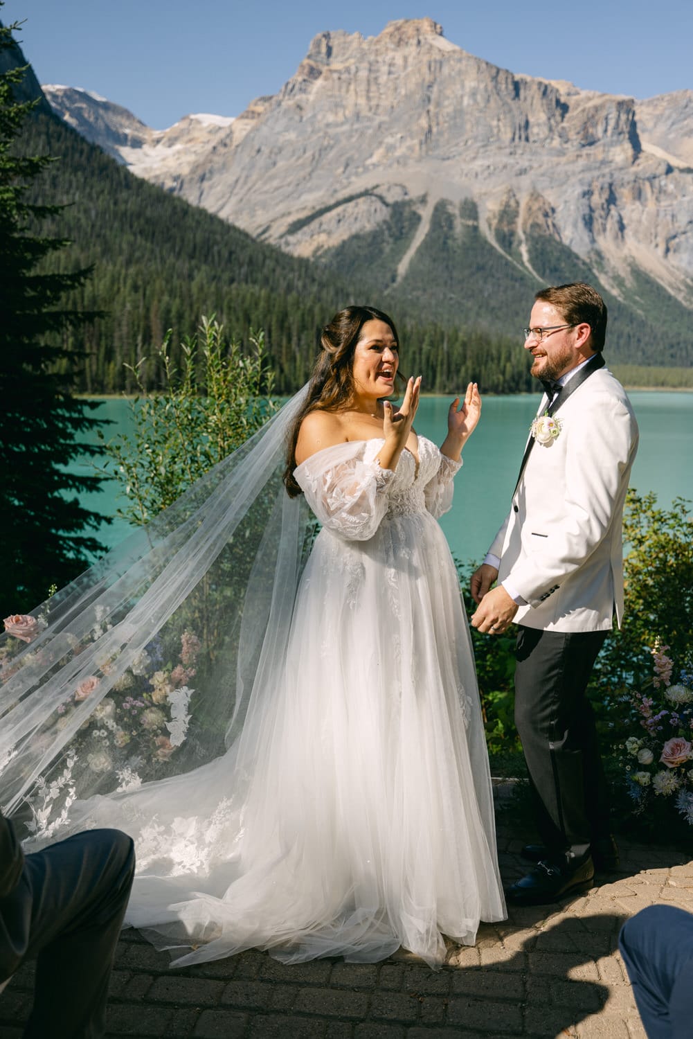 Bride in a white dress with a long veil and groom in a suit smiling at each other by a turquoise lake with mountains in the background.