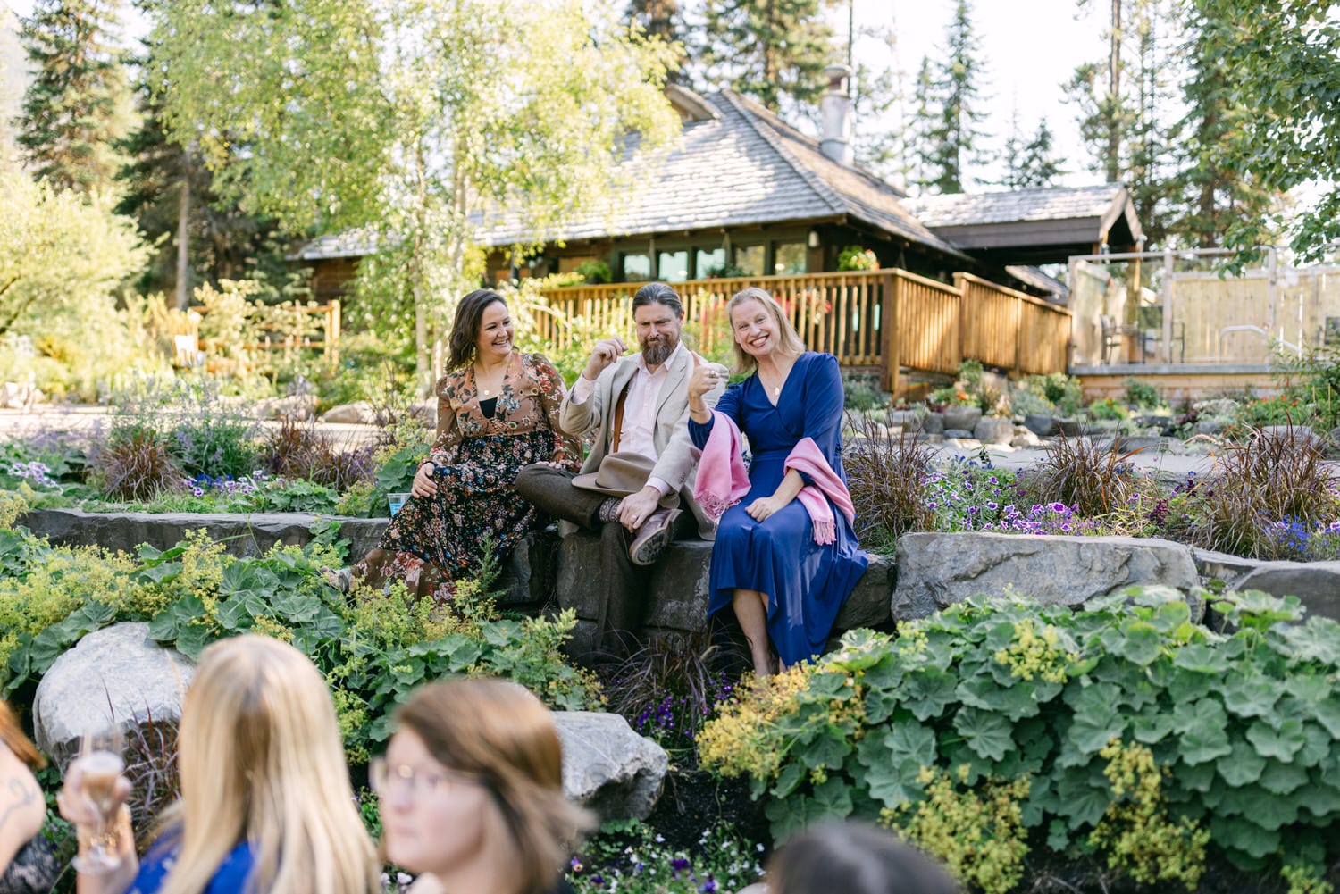 Three adults enjoying a conversation while sitting on rocks in a lush garden setting with other guests and a wooden building in the background.