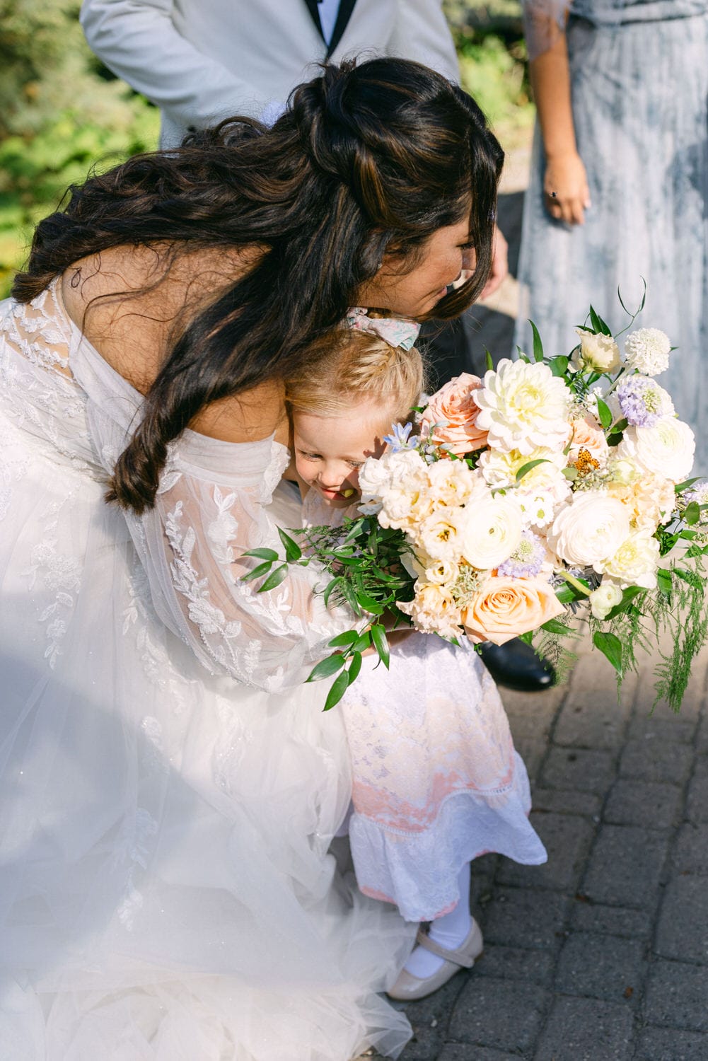 A bride in a lace wedding dress affectionately hugging a young child who is holding a flower bouquet.