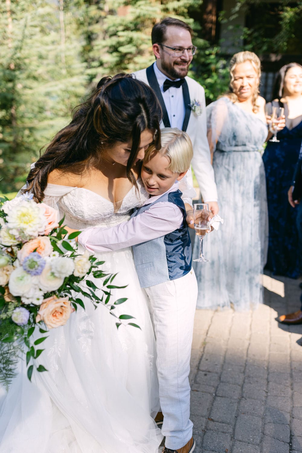 A bride in a white dress hugs a young boy in a vest at an outdoor wedding reception, with guests and greenery in the background.