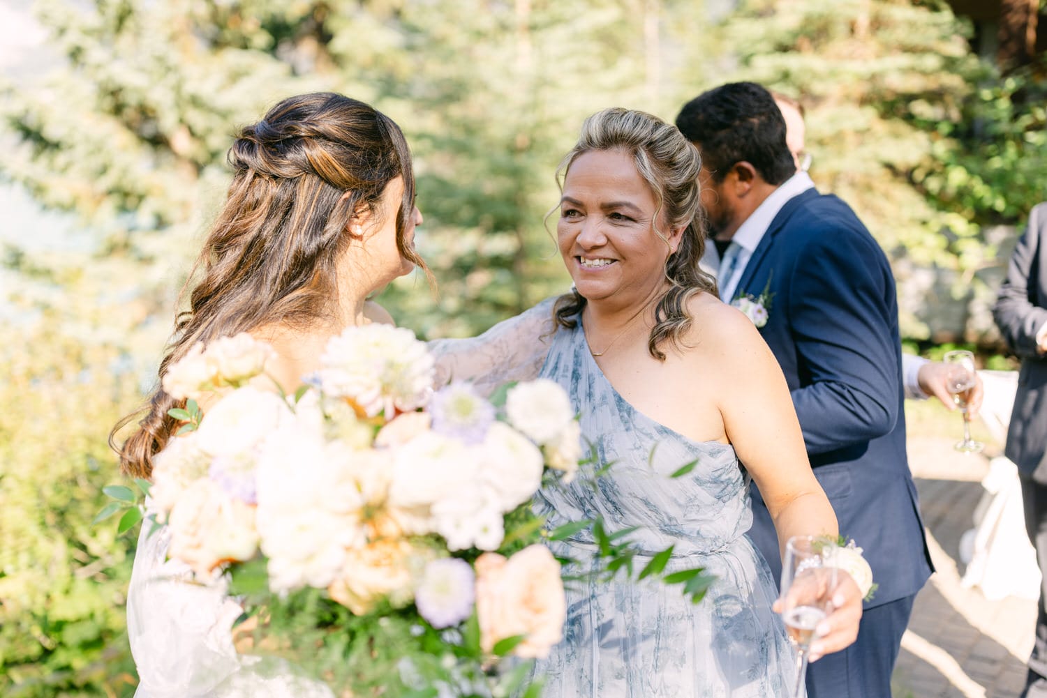 A woman in a blue dress smiling at a bride holding a bouquet, with guests and greenery in the background