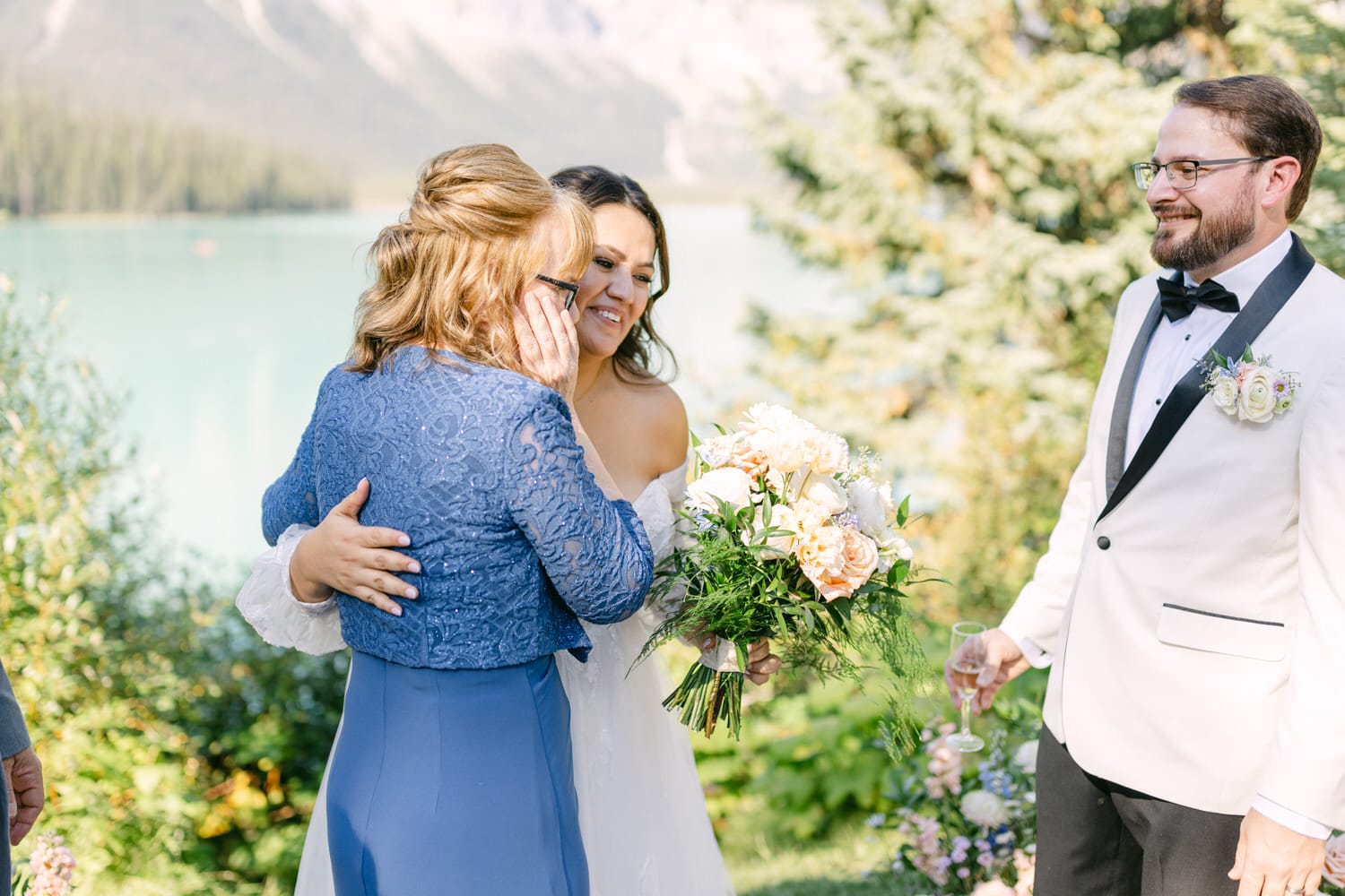 A bride embracing a woman in a blue dress while the groom looks on with a smile in an outdoor wedding setting by a lake.