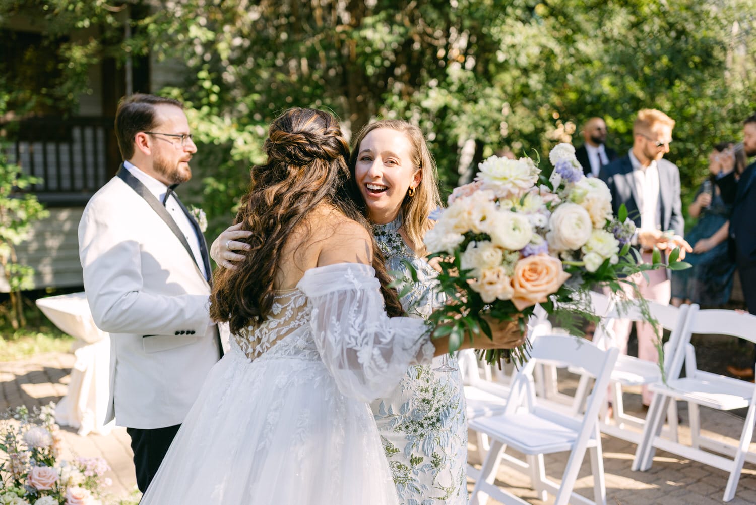 A bride in a white dress with her back turned embracing a smiling woman holding a bouquet, with wedding guests in the background.