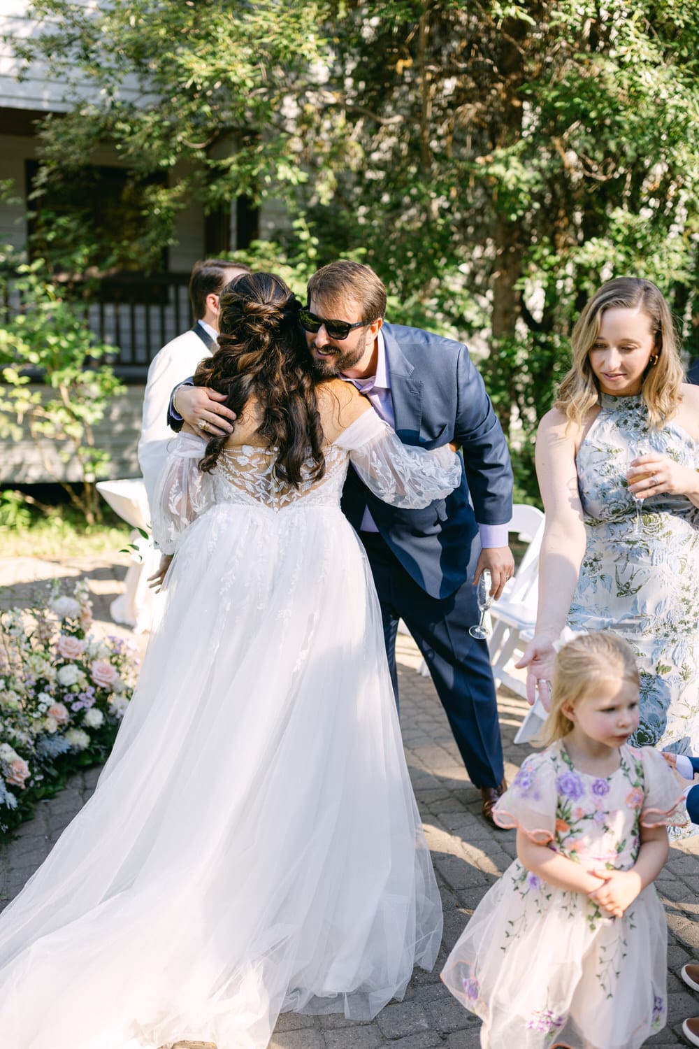 A man in a suit with sunglasses embracing a bride in a white dress at an outdoor wedding, with guests around them.