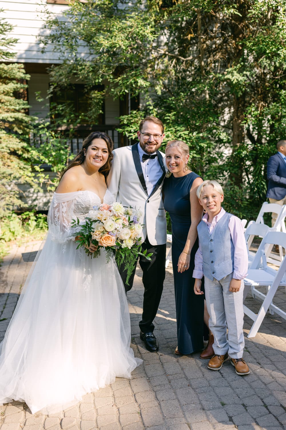 A bride and groom smiling with two family members outdoors on a sunny day, all dressed in formal attire with greenery in the background.