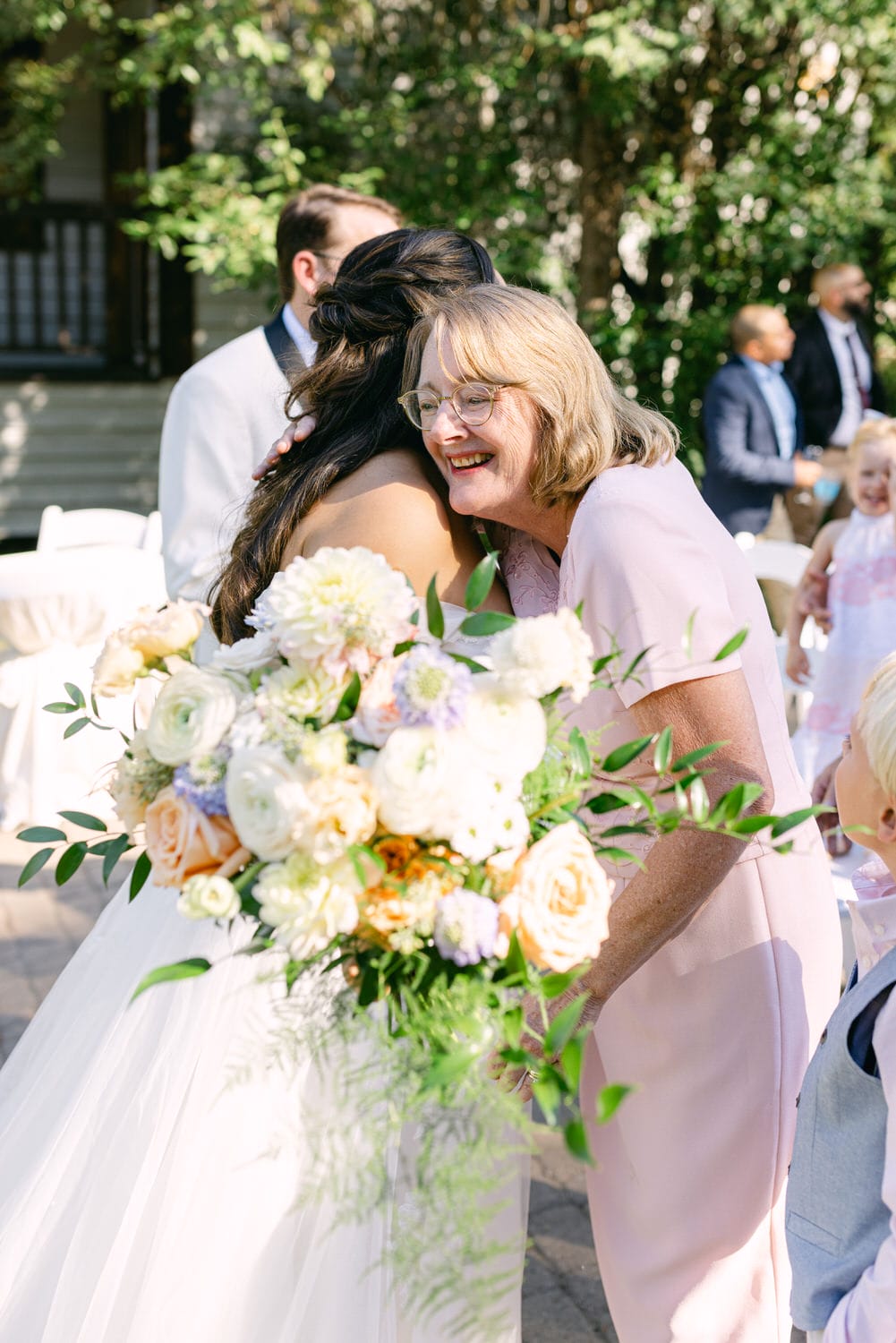 An older woman in a pink dress hugging a bride holding a bouquet, with wedding guests in the background.