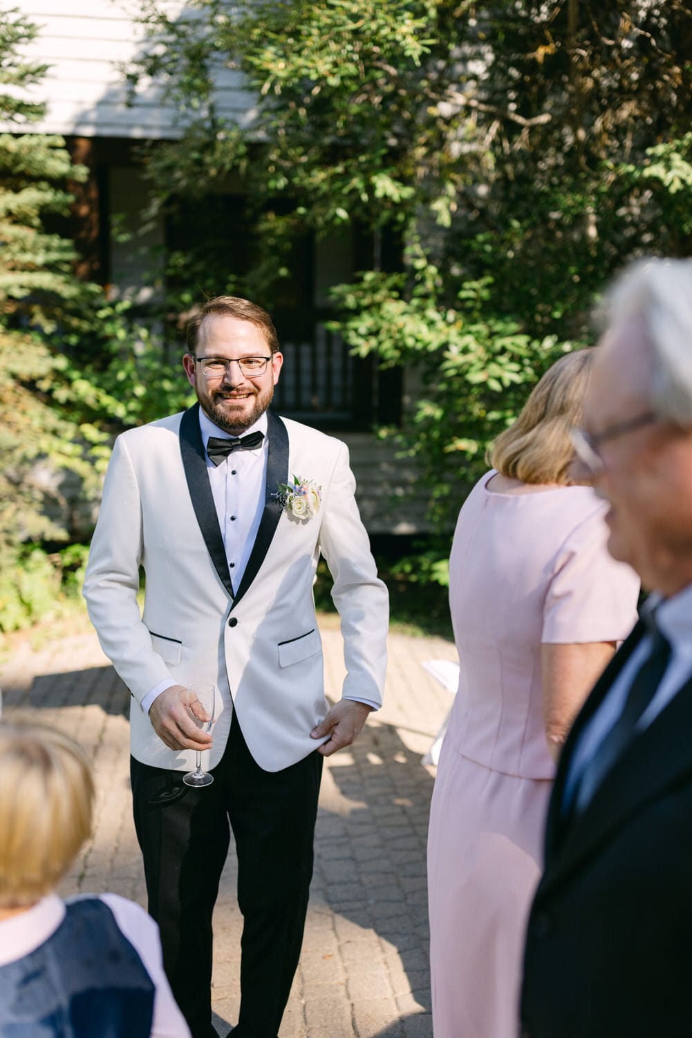 A cheerful groom in a stylish white tuxedo with a bow tie and boutonniere, holding a wine glass, at an outdoor wedding setting.