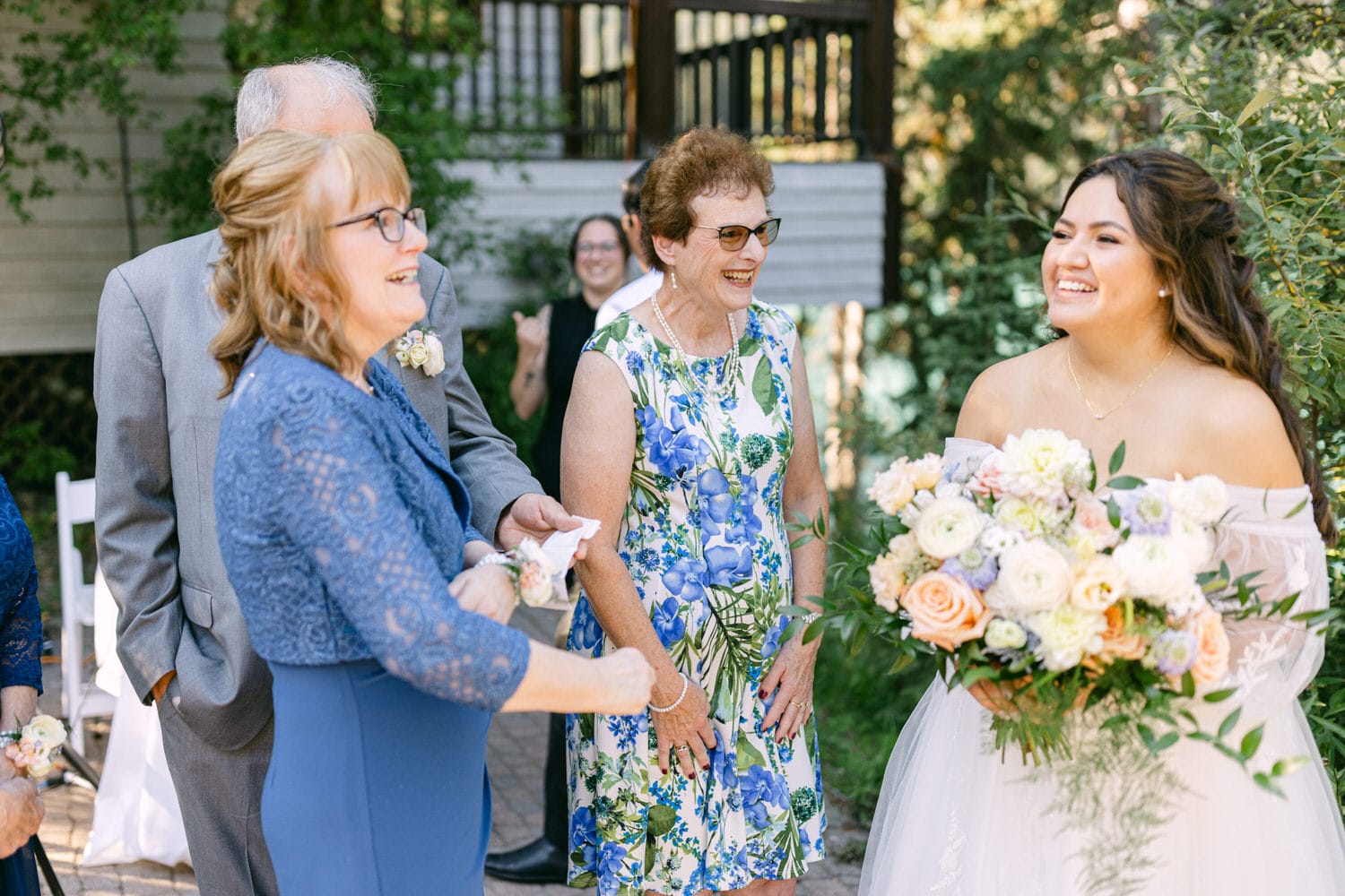 A smiling bride holding a bouquet shares a happy moment with laughing guests at an outdoor wedding ceremony.