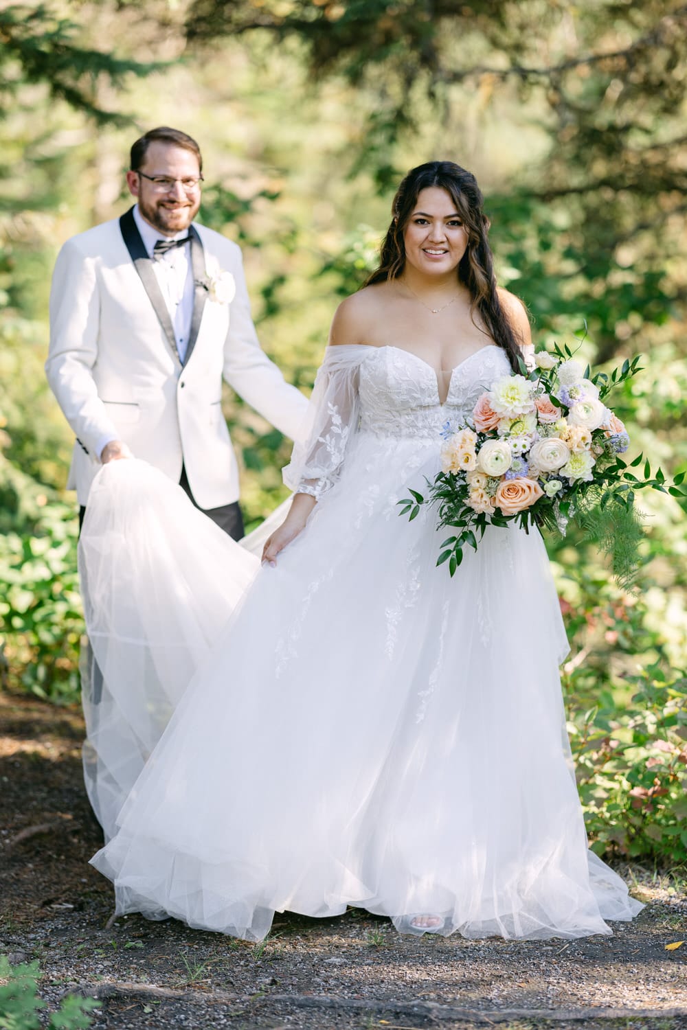 A bride in a white off-shoulder wedding dress holding a bouquet and a groom in a white suit smiling in a sunlit forest setting.