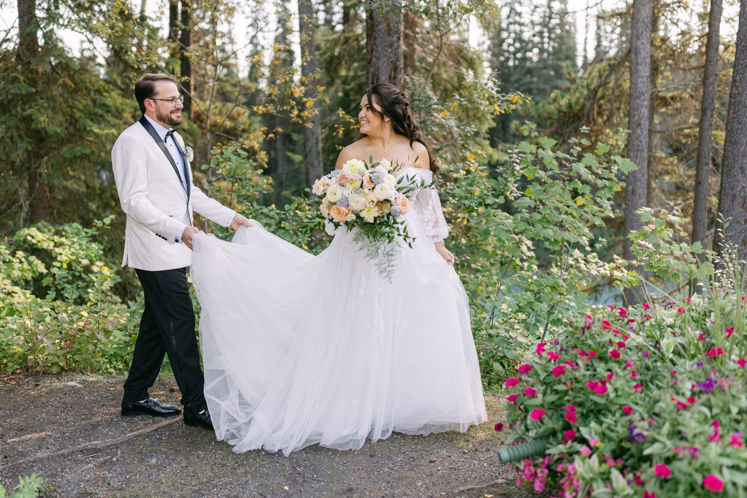 A bride and groom smiling at each other in a forest setting with the groom holding the bride's dress and the bride holding a bouquet of flowers.