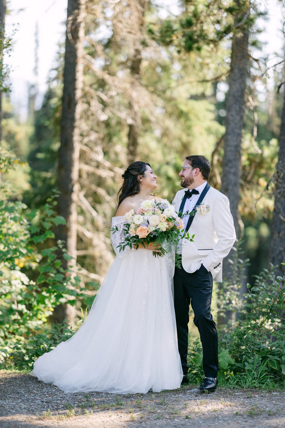 A bride and groom in wedding attire smiling at each other surrounded by trees.