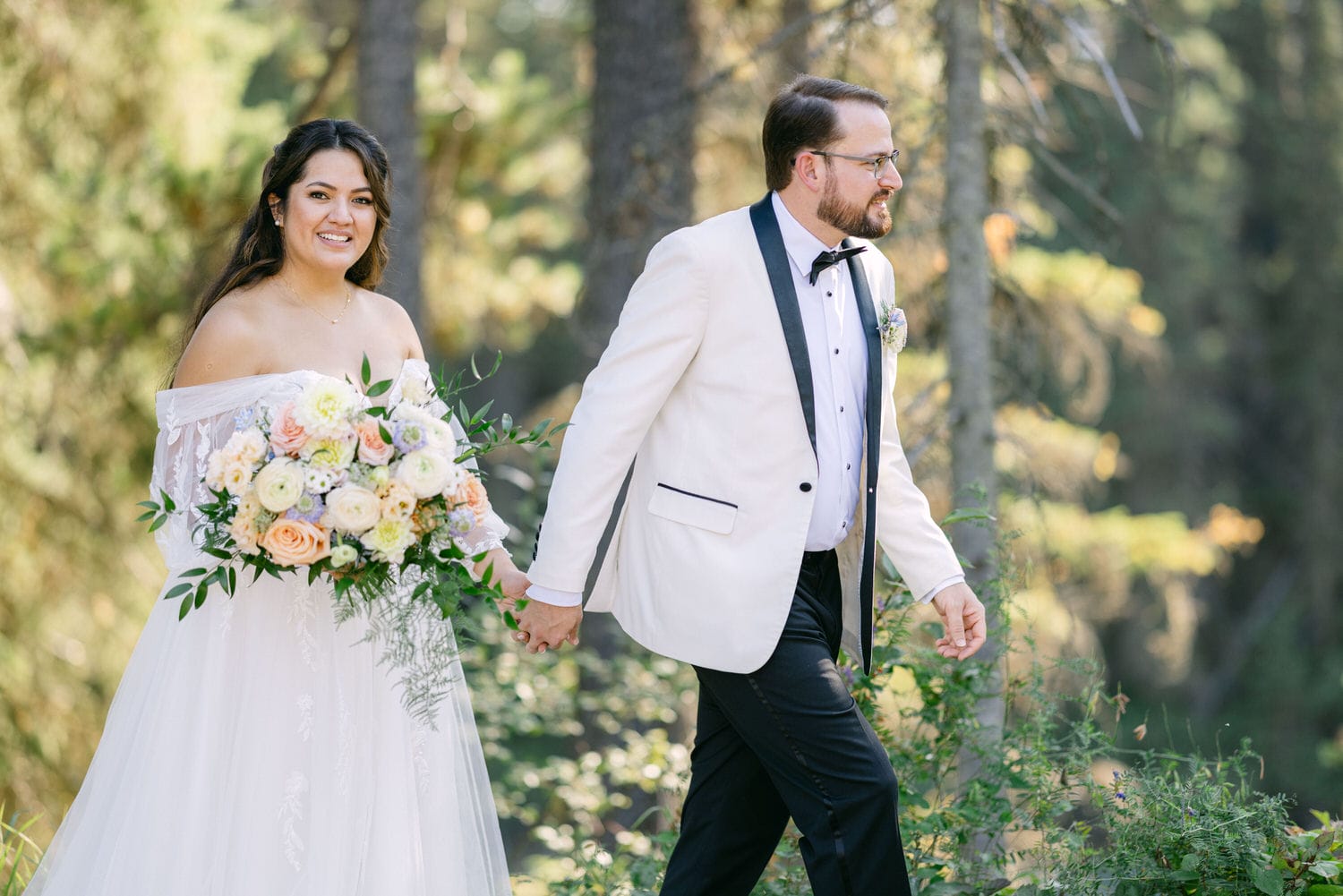 A bride and groom holding hands and walking through a forest setting, both smiling and dressed in wedding attire.