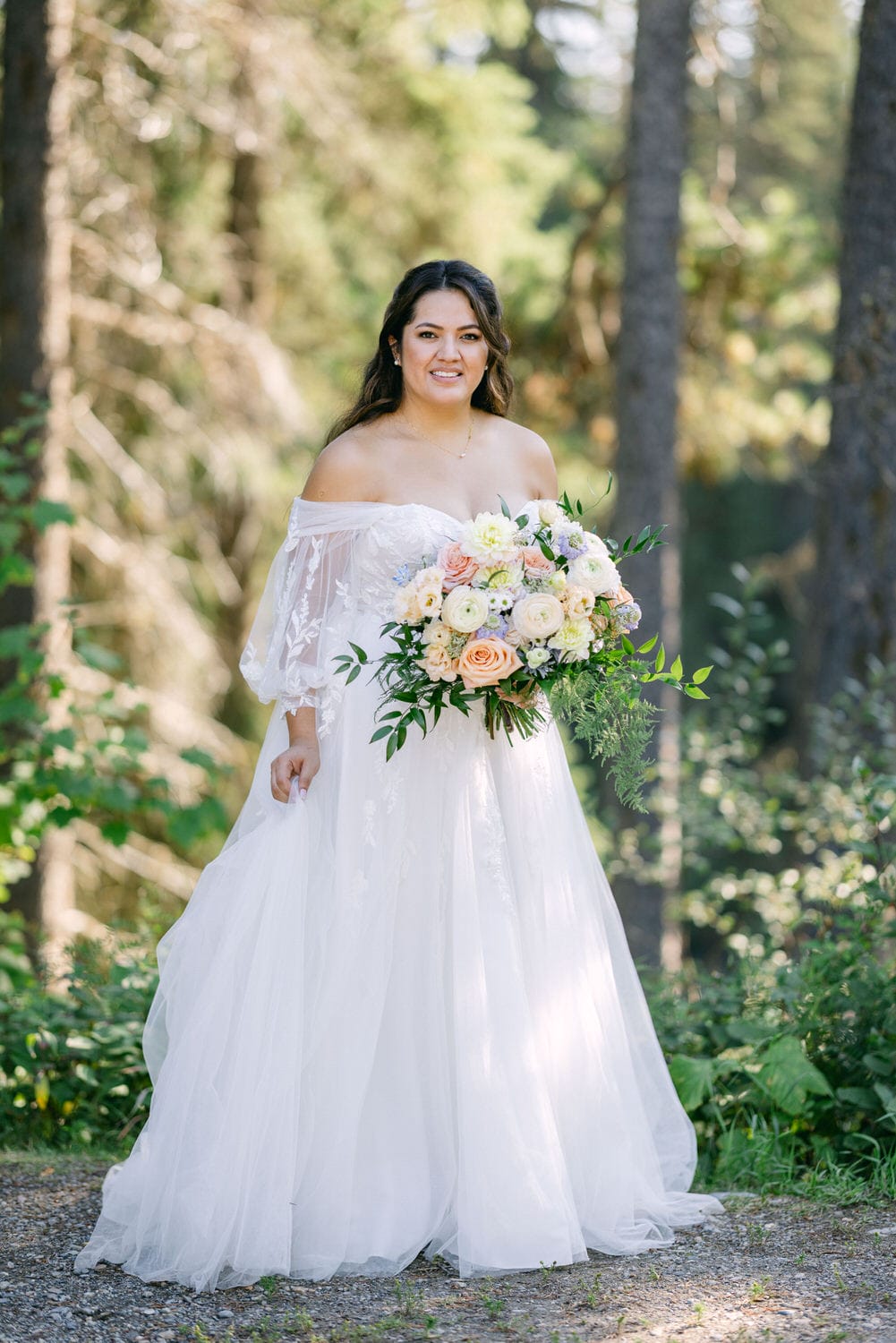 A woman in a white off-shoulder wedding dress smiling and holding a bouquet of flowers in a forest setting.