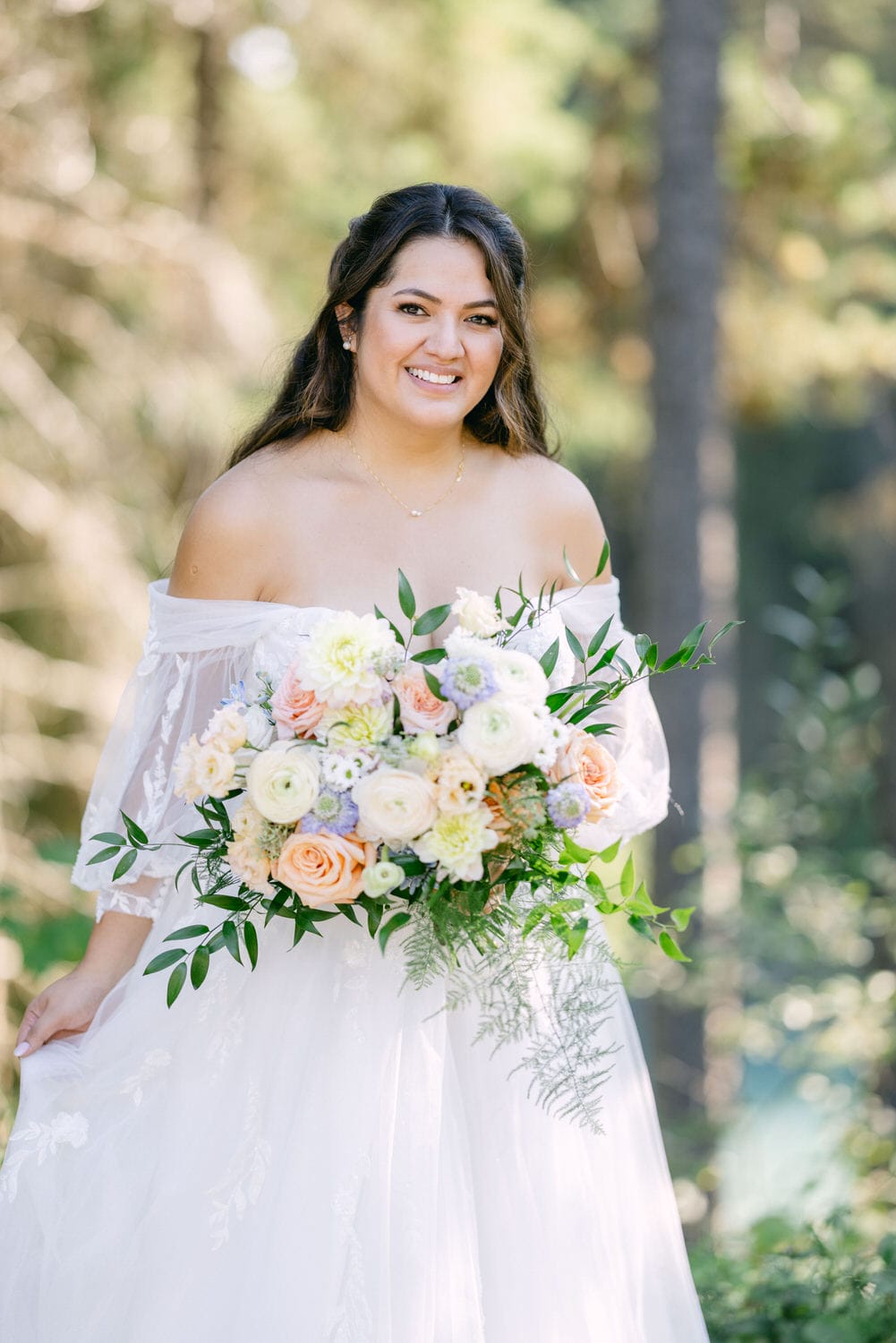 A smiling bride in an off-shoulder wedding dress holding a lush bouquet of flowers outdoors.