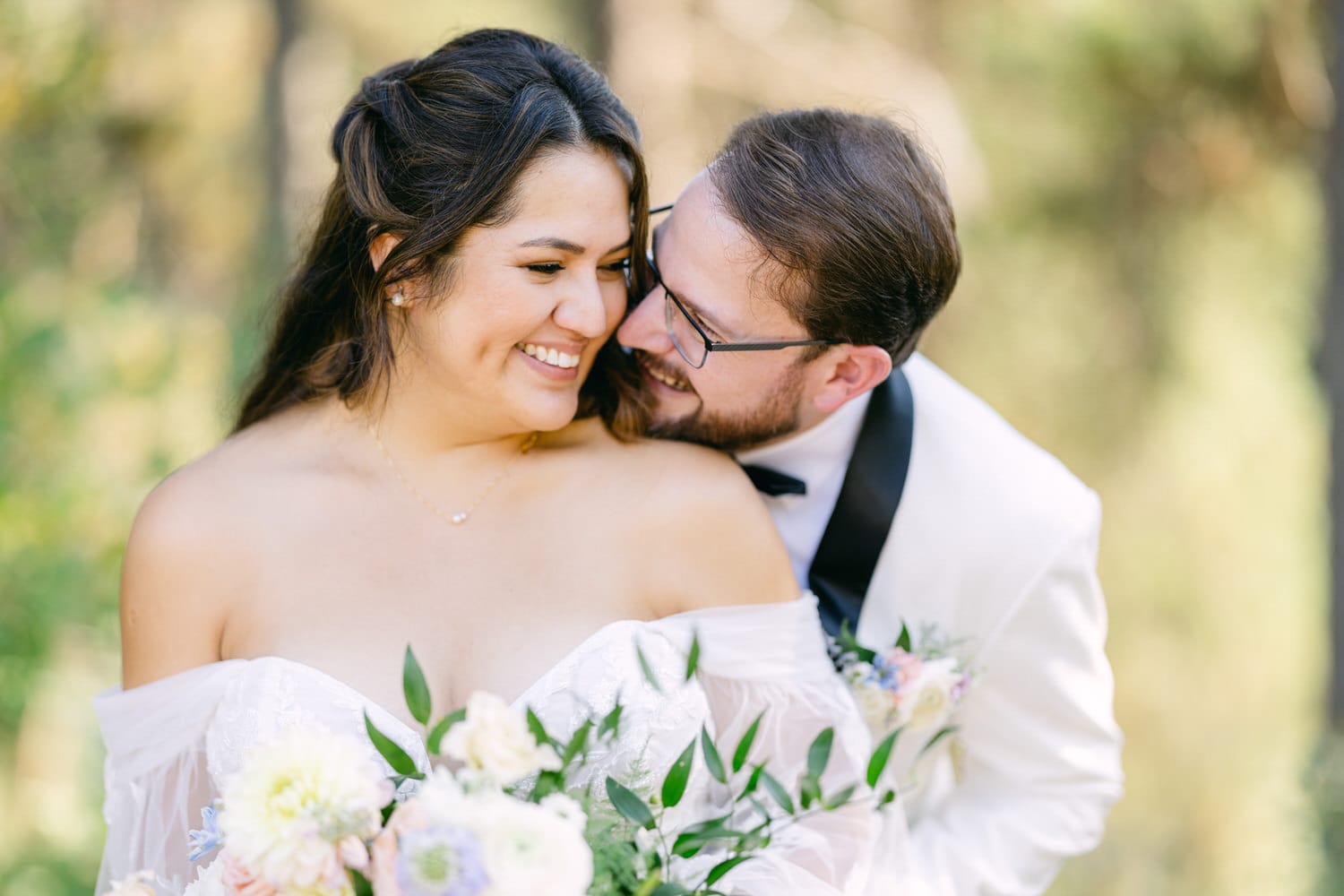 A joyful bride and groom embracing outdoors, with the groom whispering into the bride's ear while she smiles, holding a bouquet.