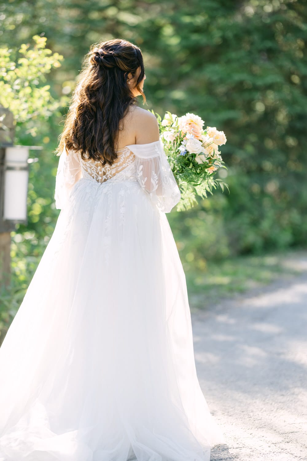 A bride in a white wedding gown holding a bouquet of flowers, captured from behind on a sunlit path.