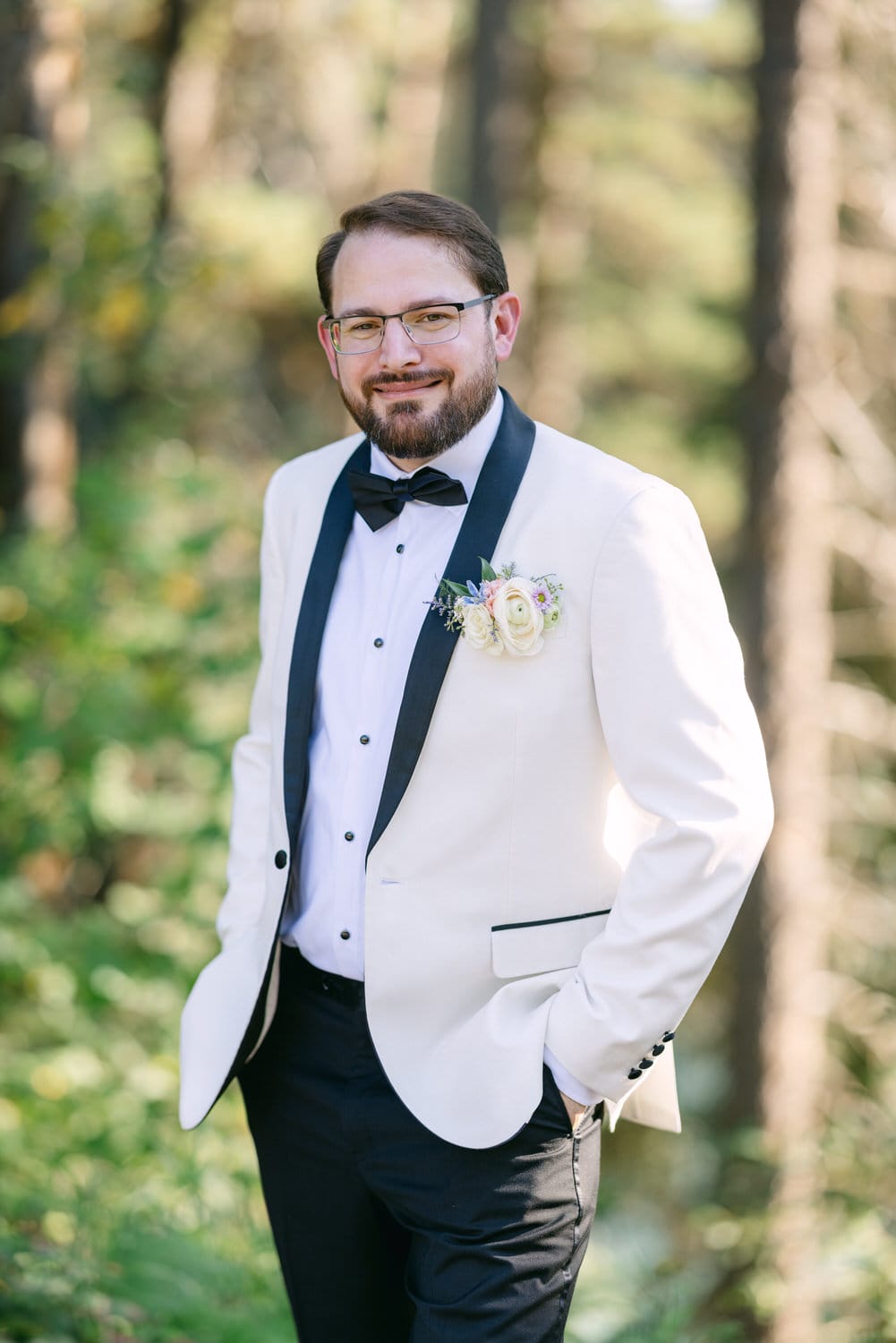 A man in a white tuxedo with a black bow tie and a floral boutonniere, standing in a forest.