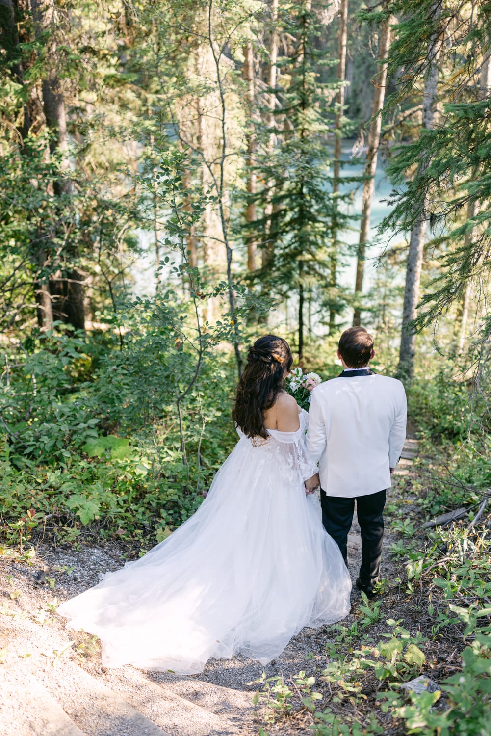 A bride and groom holding hands while walking down a forested path, with the bride in a flowing white gown and the groom in a white jacket.