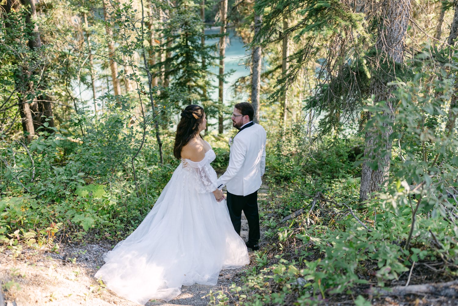 A bride and groom holding hands, walking through a lush forest with a hint of a turquoise lake in the background.