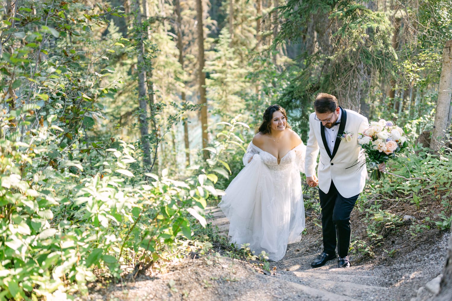 A bride and groom holding hands and smiling while walking along a forest path.