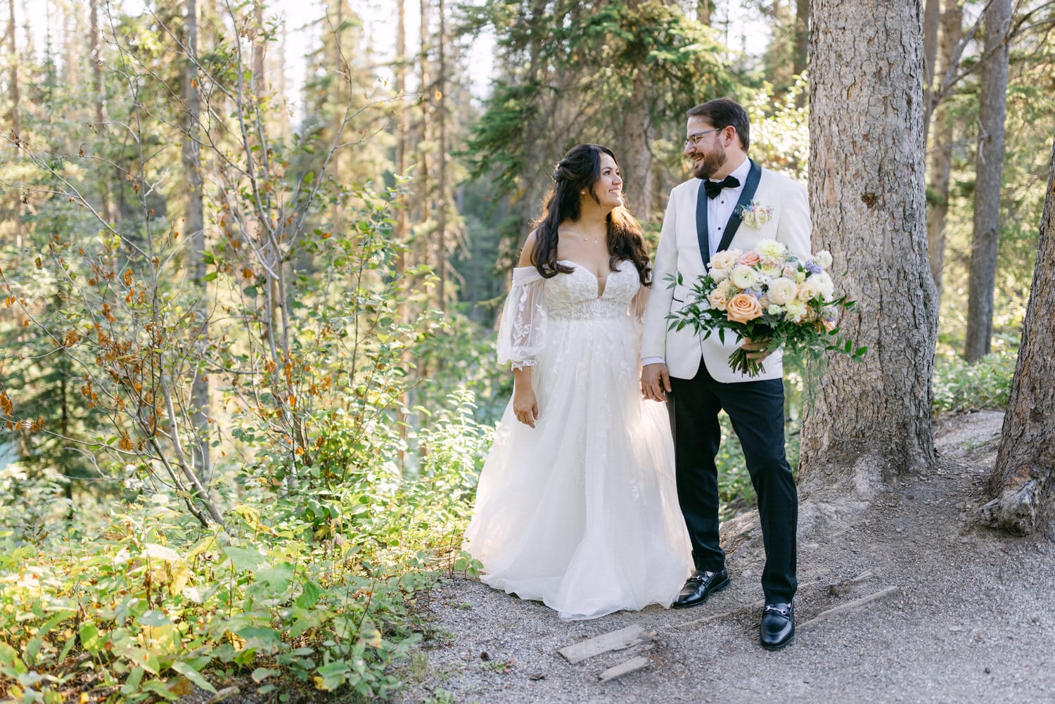 A newlywed couple holding hands and smiling at each other in a forest setting, with the bride holding a bouquet of flowers.