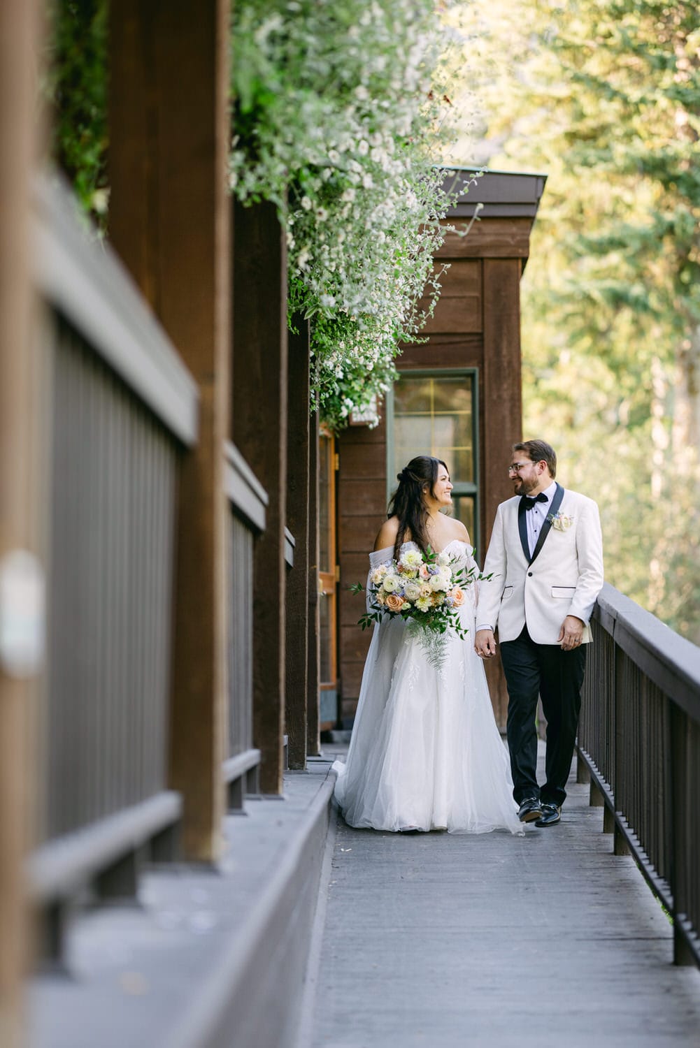 A bride and groom walking hand in hand along a wooden deck adorned with hanging white flowers, with a backdrop of lush greenery.