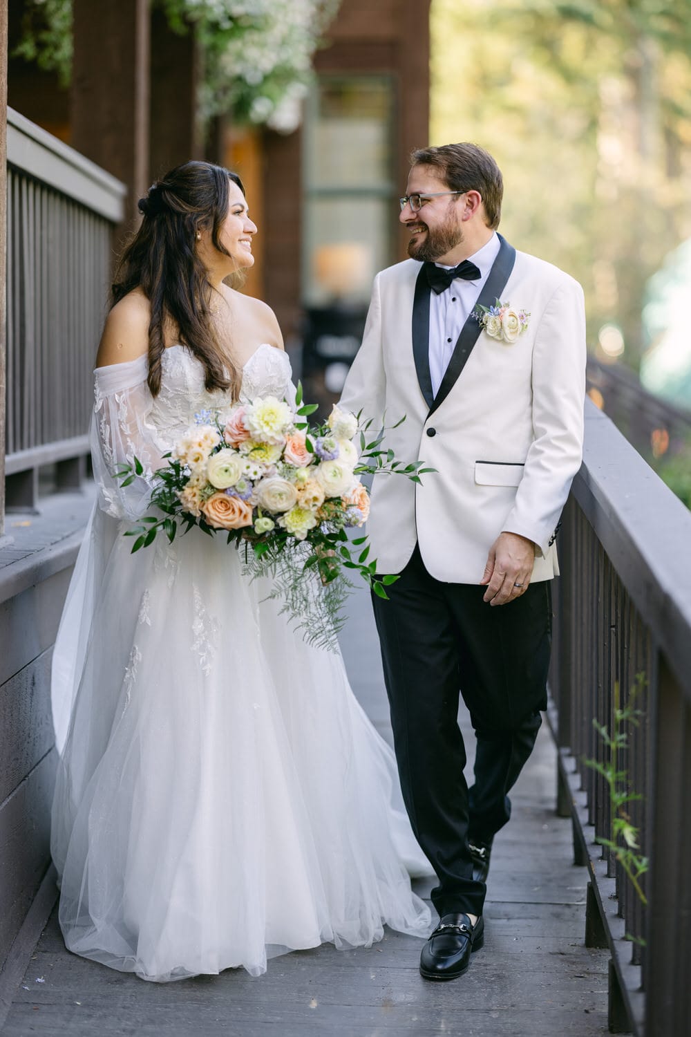 A bride and groom sharing a smile while walking on a wooden bridge, the bride holding a bouquet and both dressed in wedding attire.