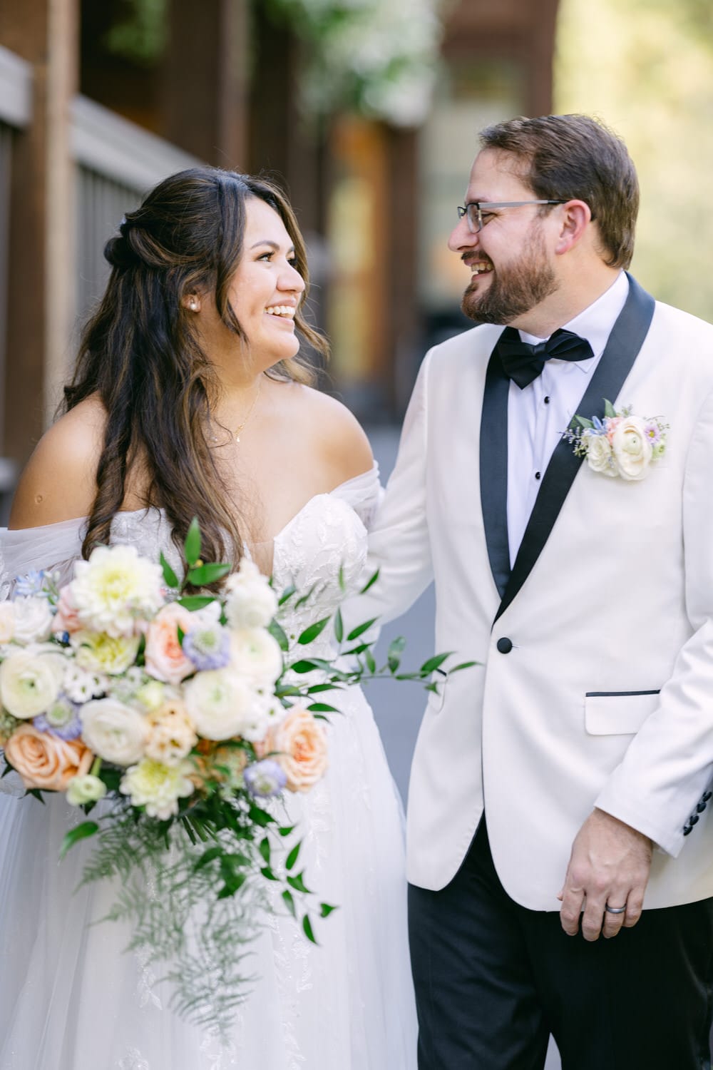 A bride and groom smiling at each other, the bride holding a bouquet of flowers, both dressed in wedding attire.