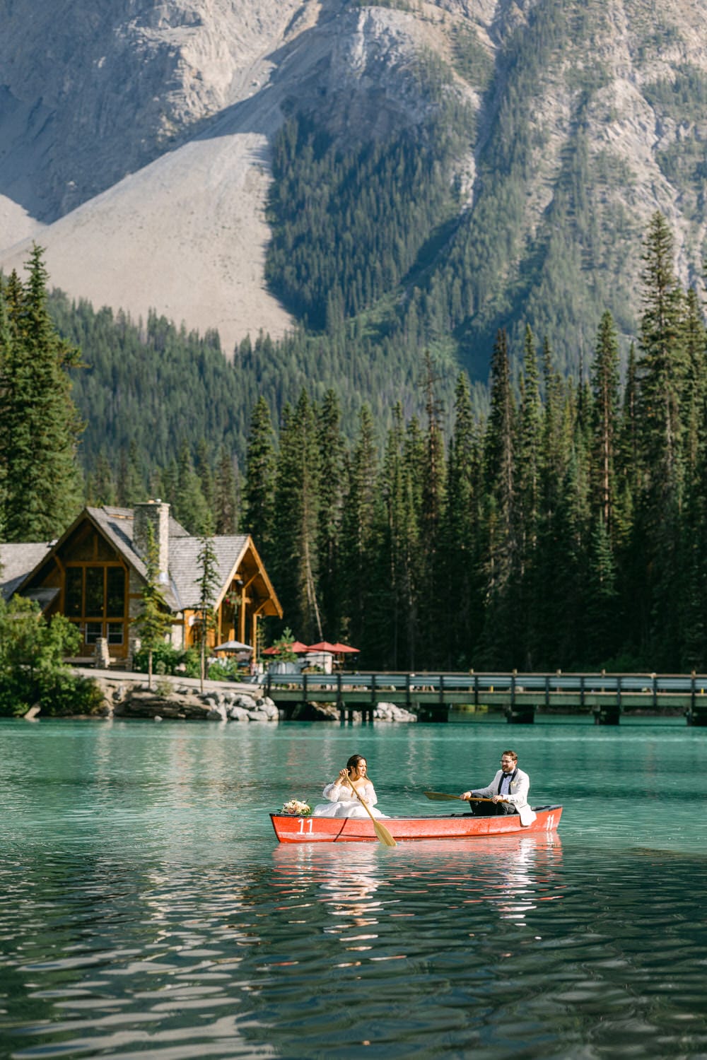 A couple in formal attire canoeing on a serene mountain lake with a log cabin and pine trees in the background.