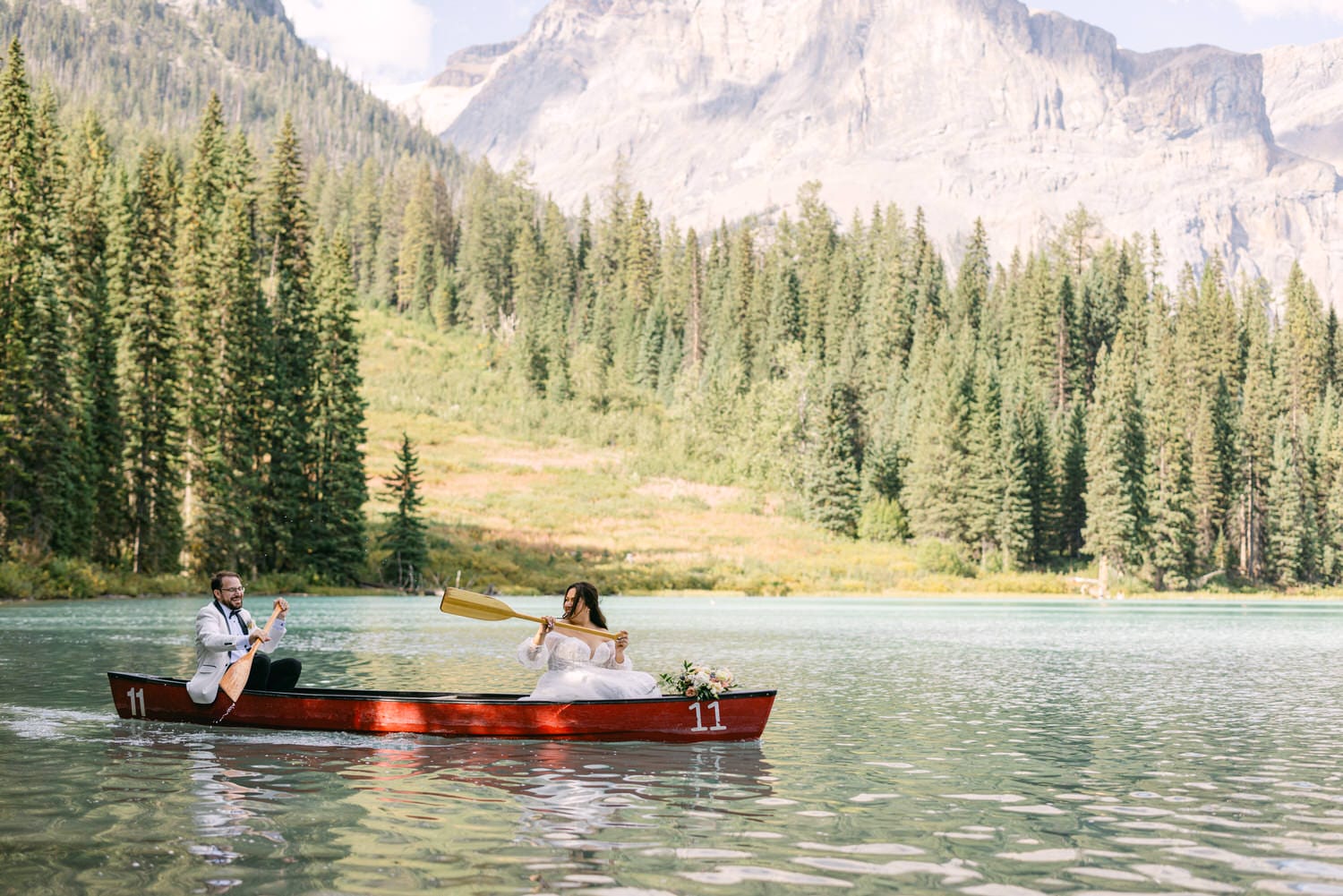 A couple in wedding attire paddling a canoe on a serene lake with mountainous and forested backdrop.