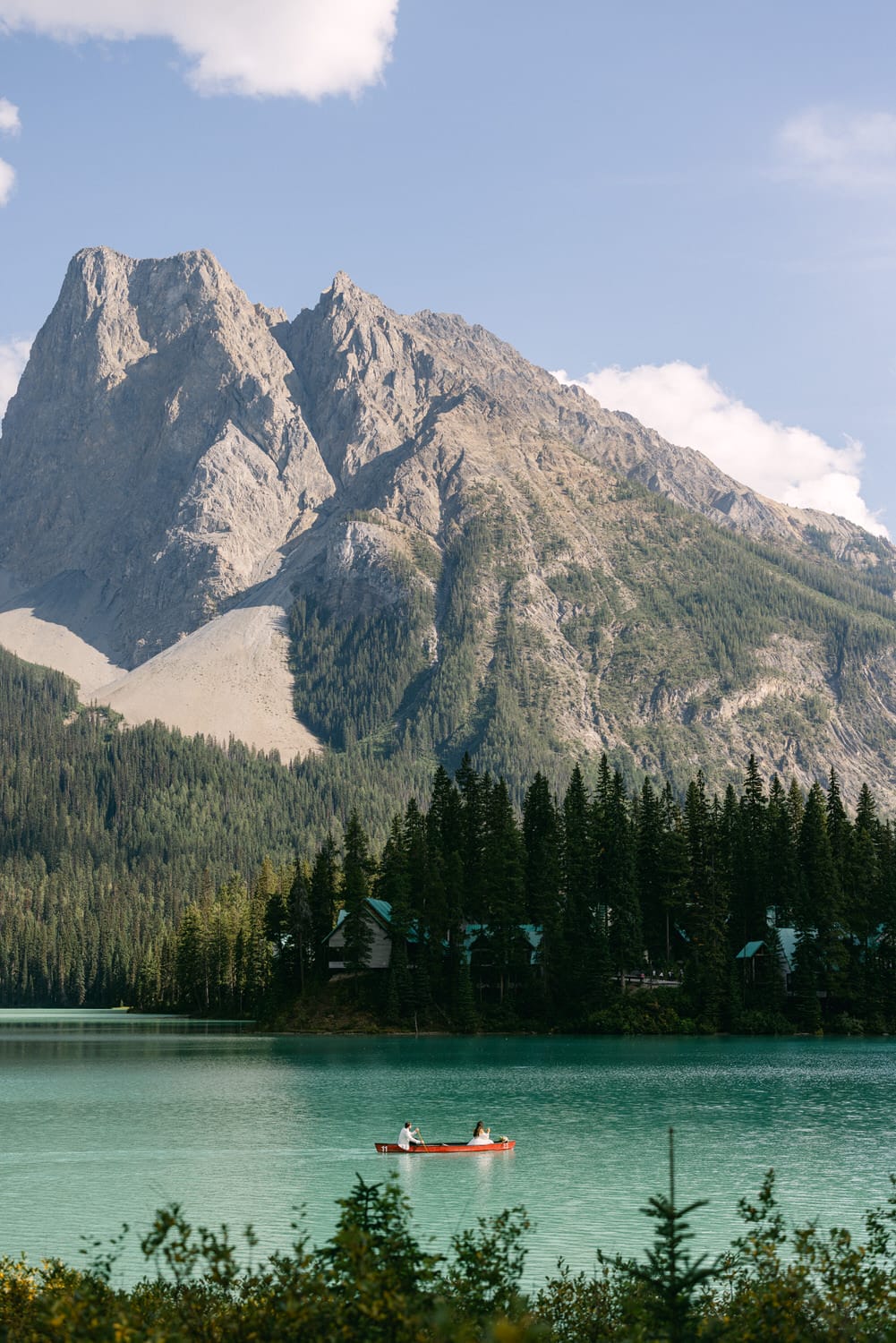 Two people canoeing on a calm turquoise lake with towering mountains in the background and evergreen trees lining the shore.