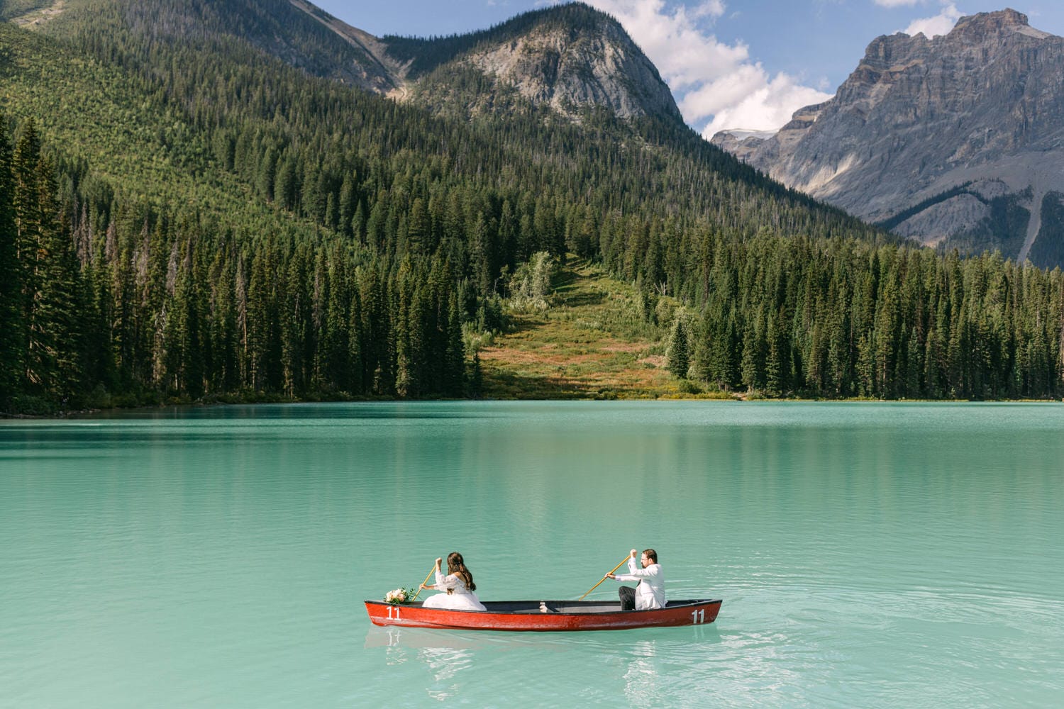 A couple paddling a red canoe on a tranquil turquoise lake with lush green forests and mountains in the background.