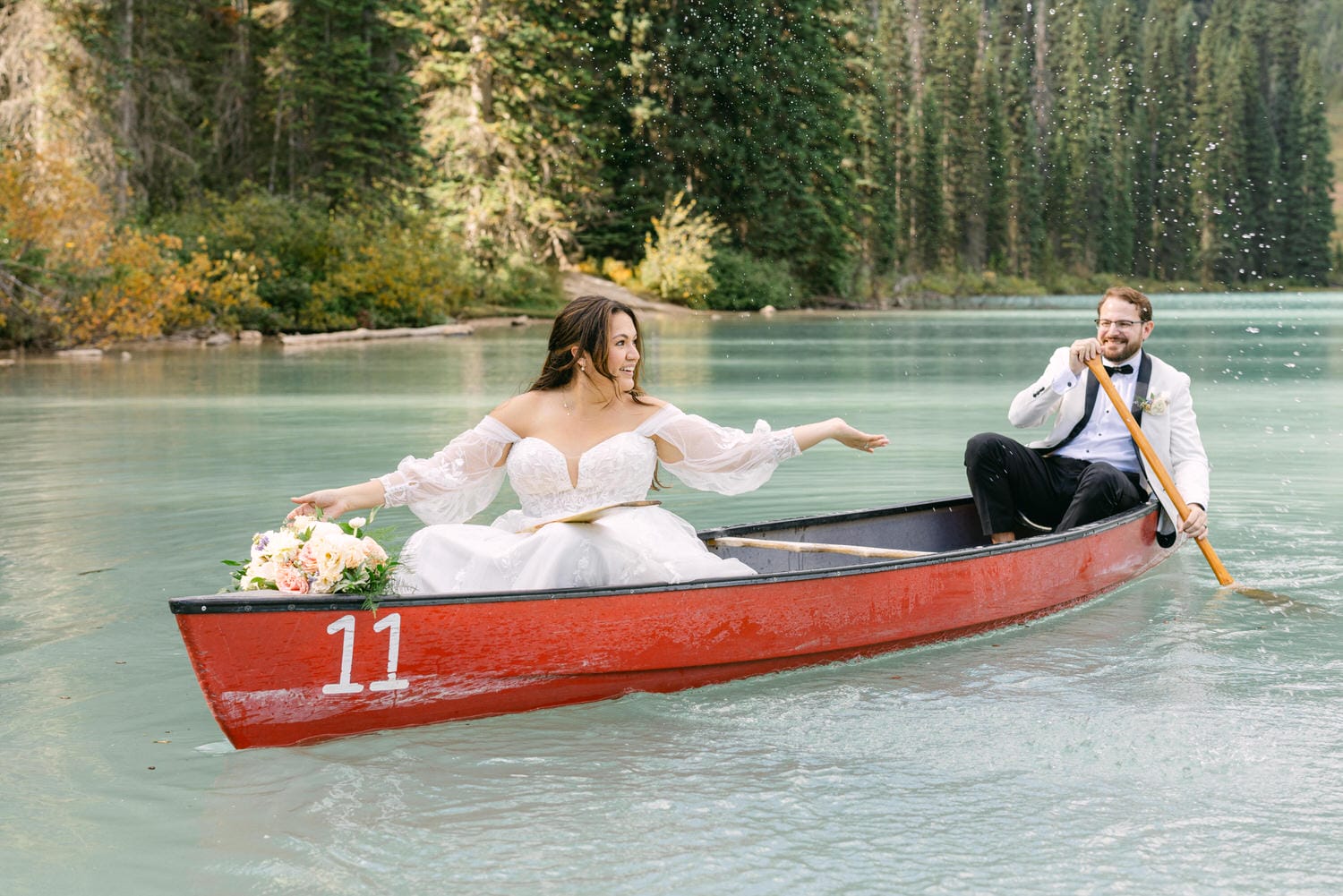 A bride and groom in wedding attire joyfully rowing in a red canoe on a scenic lake with greenery in the background.