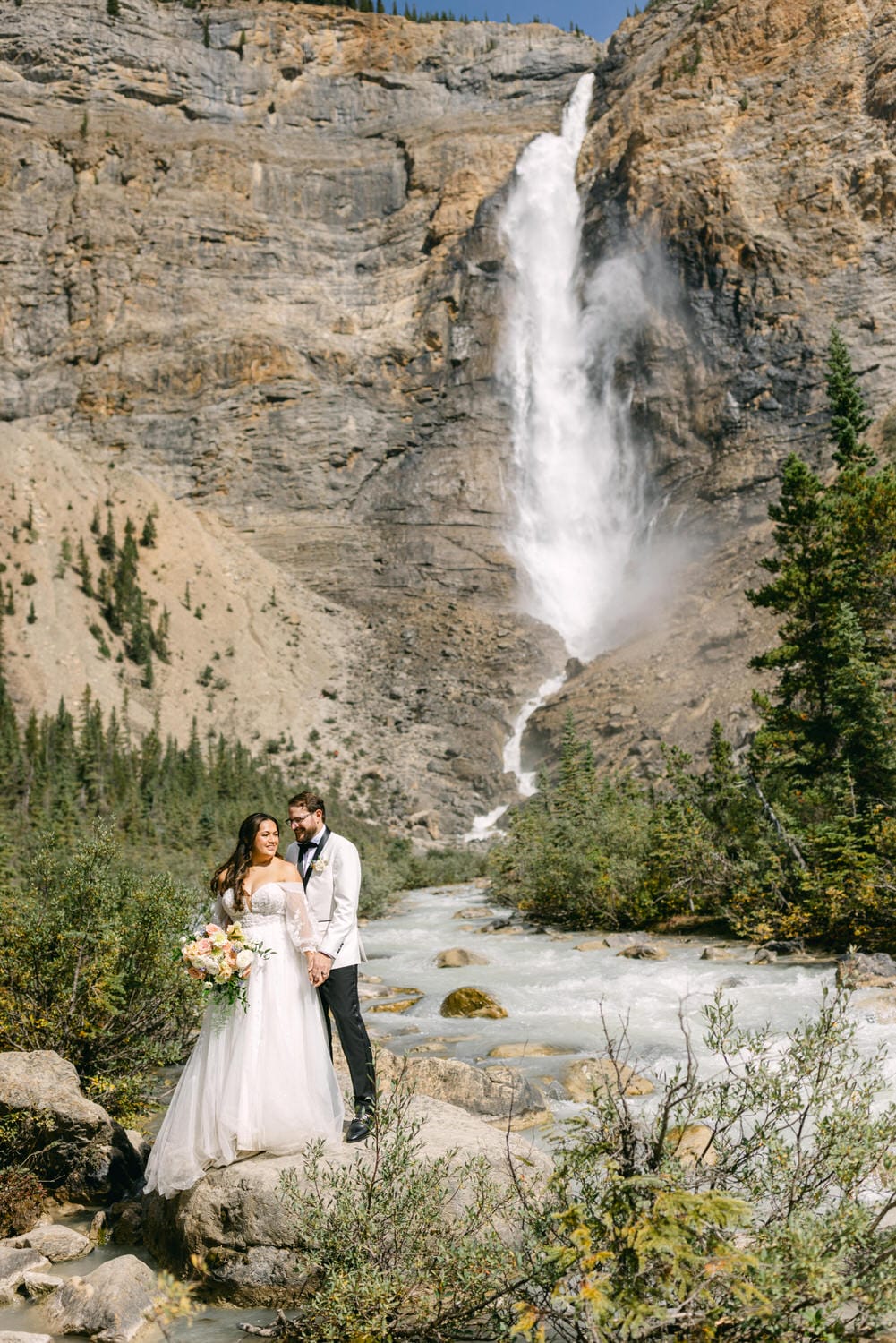 A bride and groom holding hands in front of a majestic waterfall with a flowing river surrounded by forested cliffs.