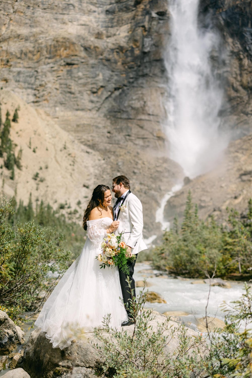 A bride and groom embracing in front of a cascading waterfall amidst mountain scenery.