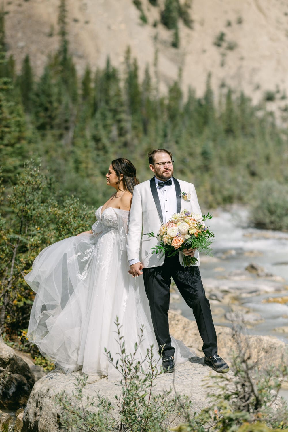 A bride and groom standing on a rocky riverbank in a mountainous landscape, with the groom holding a bouquet and looking forward while the bride looks to the side.