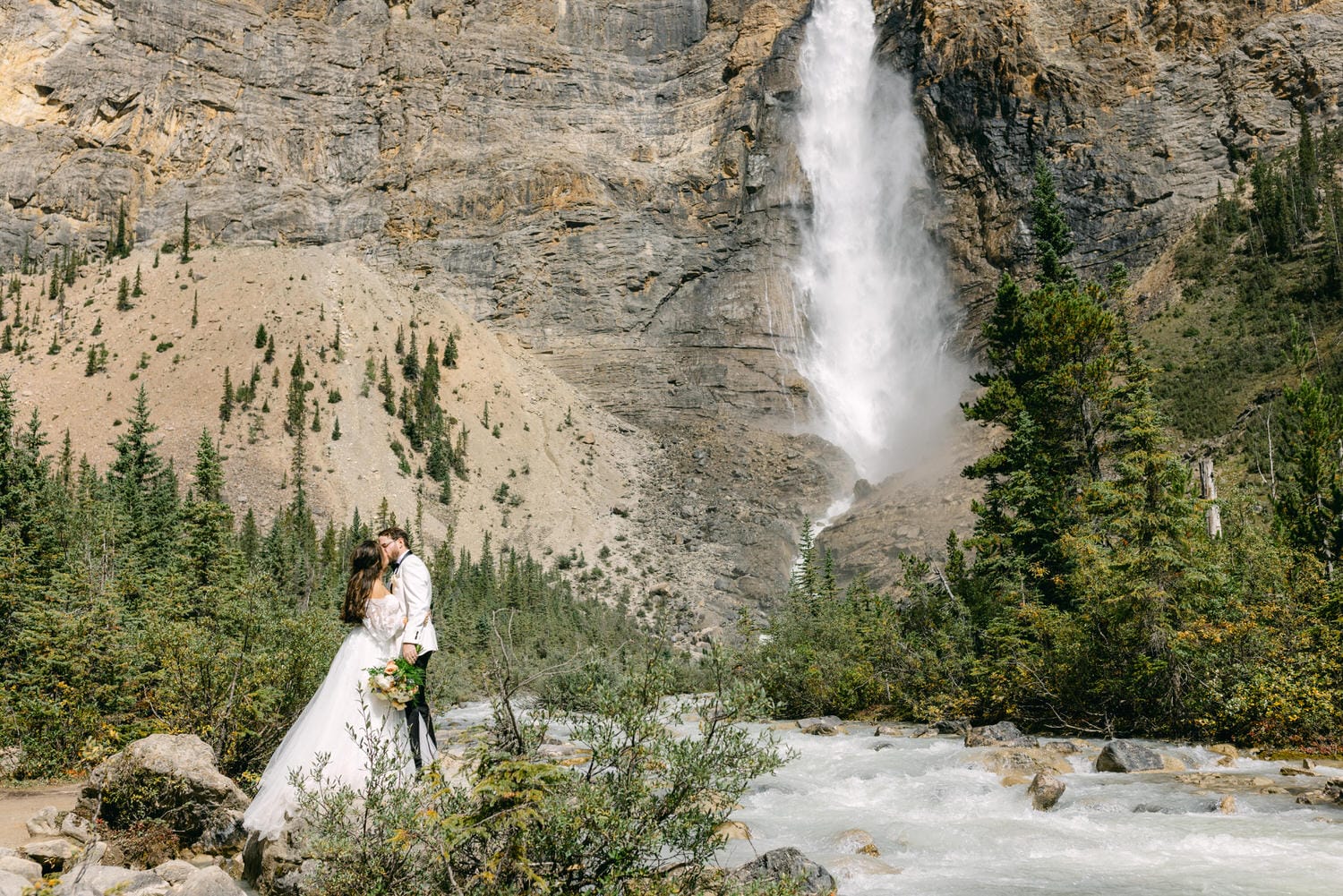 A couple in wedding attire kissing by a river with a tall waterfall and forested cliffs in the background