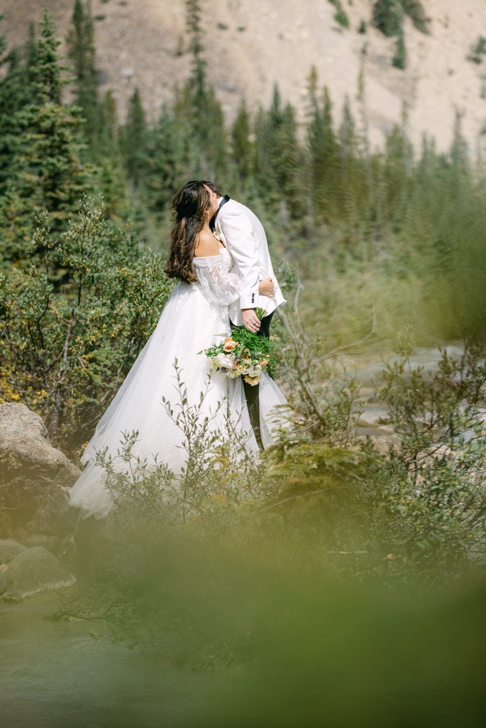 A bride in a white dress with a bouquet of flowers standing in a natural setting with green foliage and a mountainous backdrop.