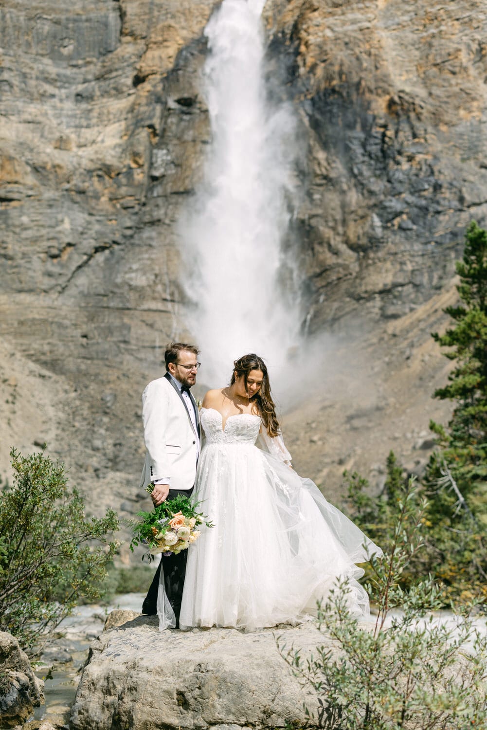 A bride and groom standing together on a rock with a large waterfall in the background.