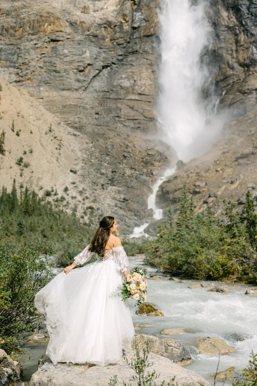 A woman in a white bridal gown stands on a rock by a river, holding a bouquet, with a waterfall in the background.