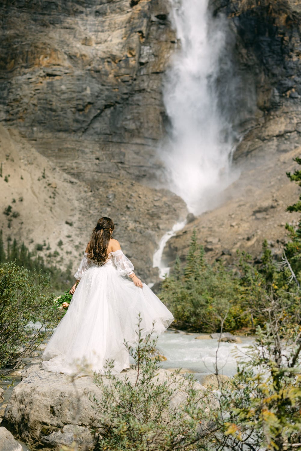 A woman in a white bridal gown standing in front of a powerful waterfall in a rocky, forested landscape.