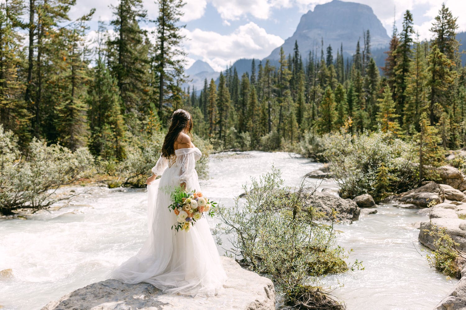 A woman in a white bridal dress holding a bouquet while standing beside a mountain stream, with forest and mountains in the background.