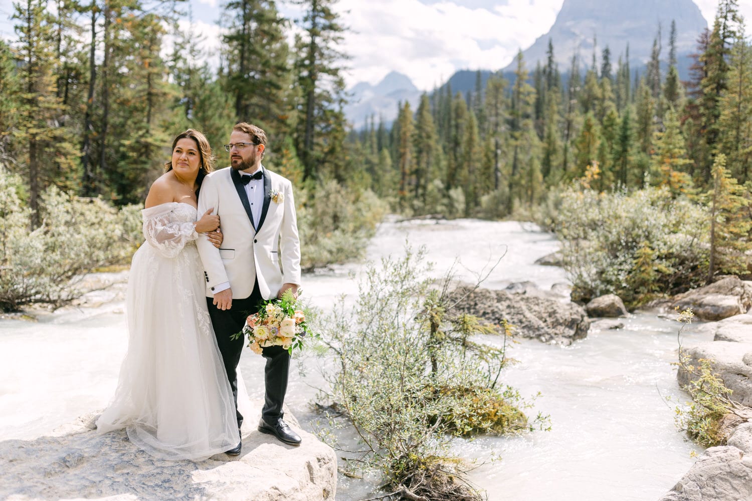 A bride and groom posing for a wedding photo by a mountain river with forest and mountains in the background.