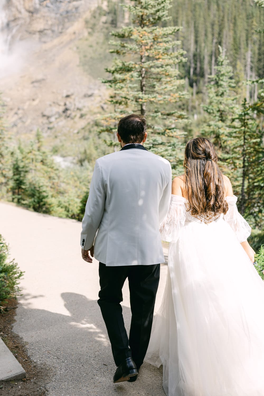 A bride and groom walking hand in hand on a path with a waterfall and trees in the background.