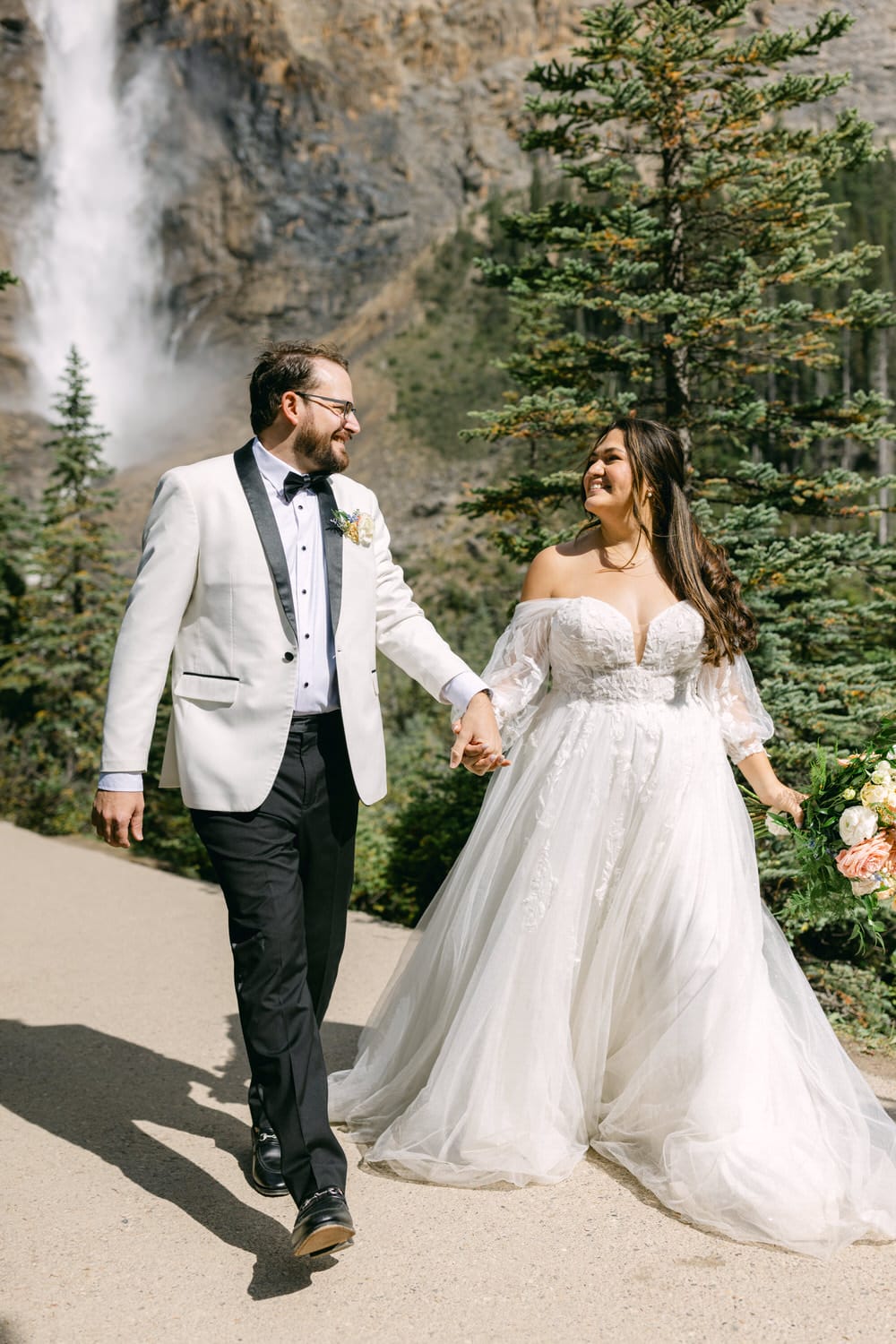 A bride and groom holding hands and smiling at each other, walking near a waterfall surrounded by greenery in daylight.