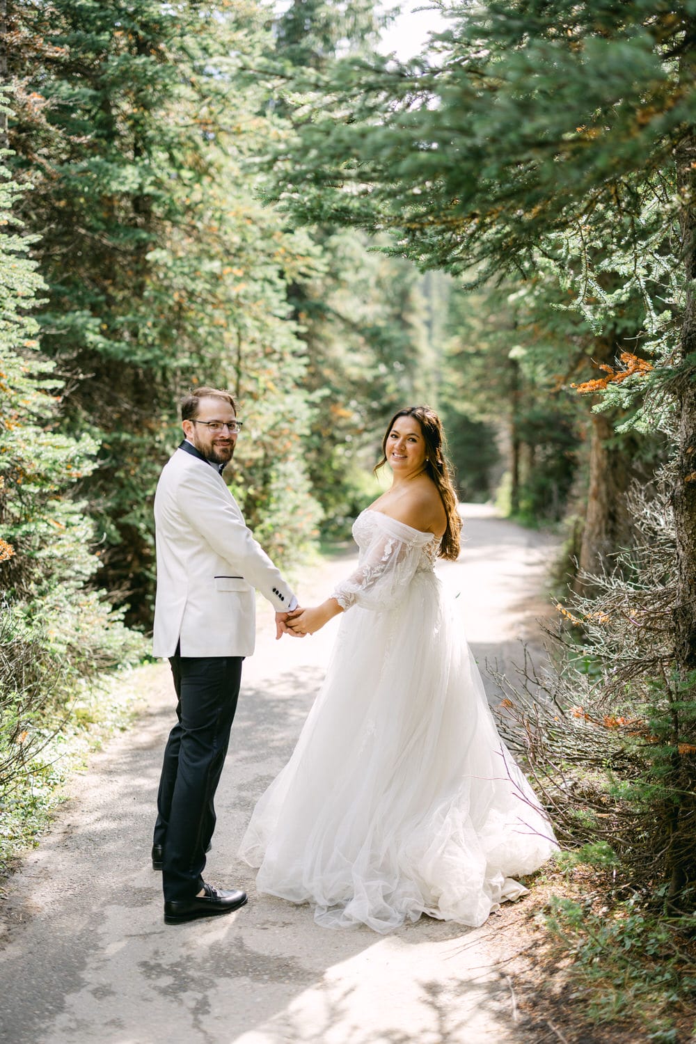 A bride and groom holding hands on a forest path, surrounded by green trees, looking back over their shoulders with smiles.