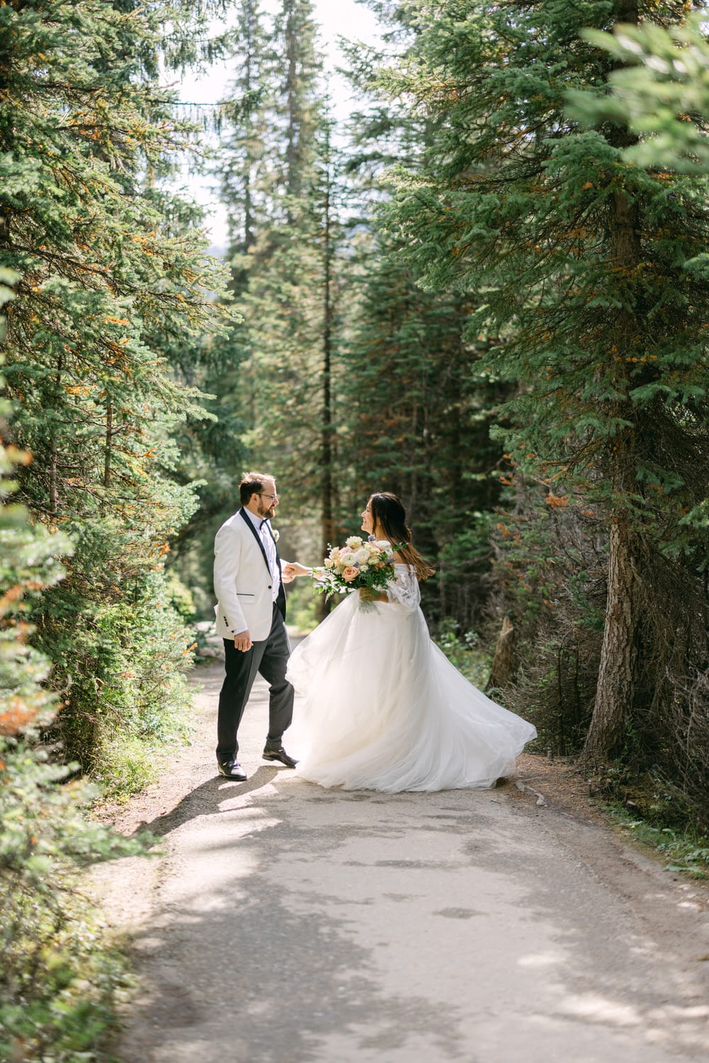 A bride in a flowing white dress and a groom in a suit holding hands on a forest path.